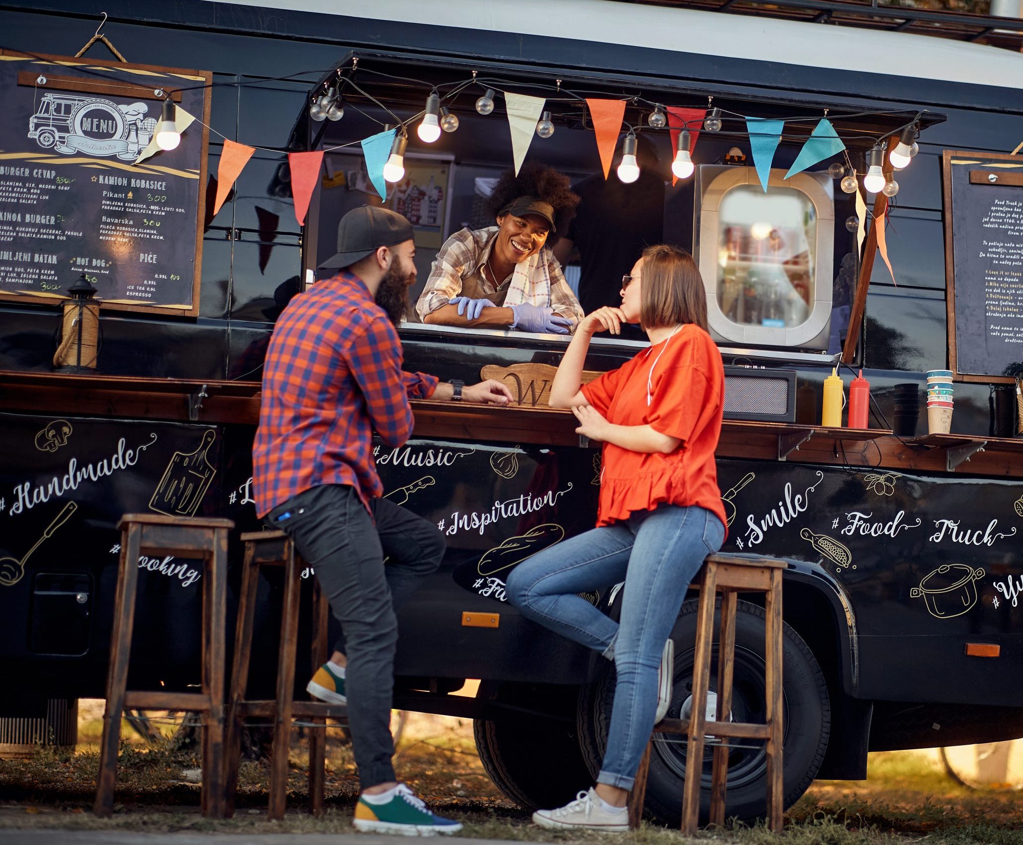 A pair of hipsters chat with the successful owner of a food truck kitted out with pennants and festoon lighting for a cozy, welcoming atmosphere.